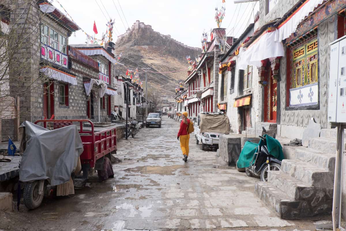 A female tourist in red jacket and yellow trousers walking down a traditional Tibetan village street with low-rise houses in mountain-backed Gyantse when travelling Tibet