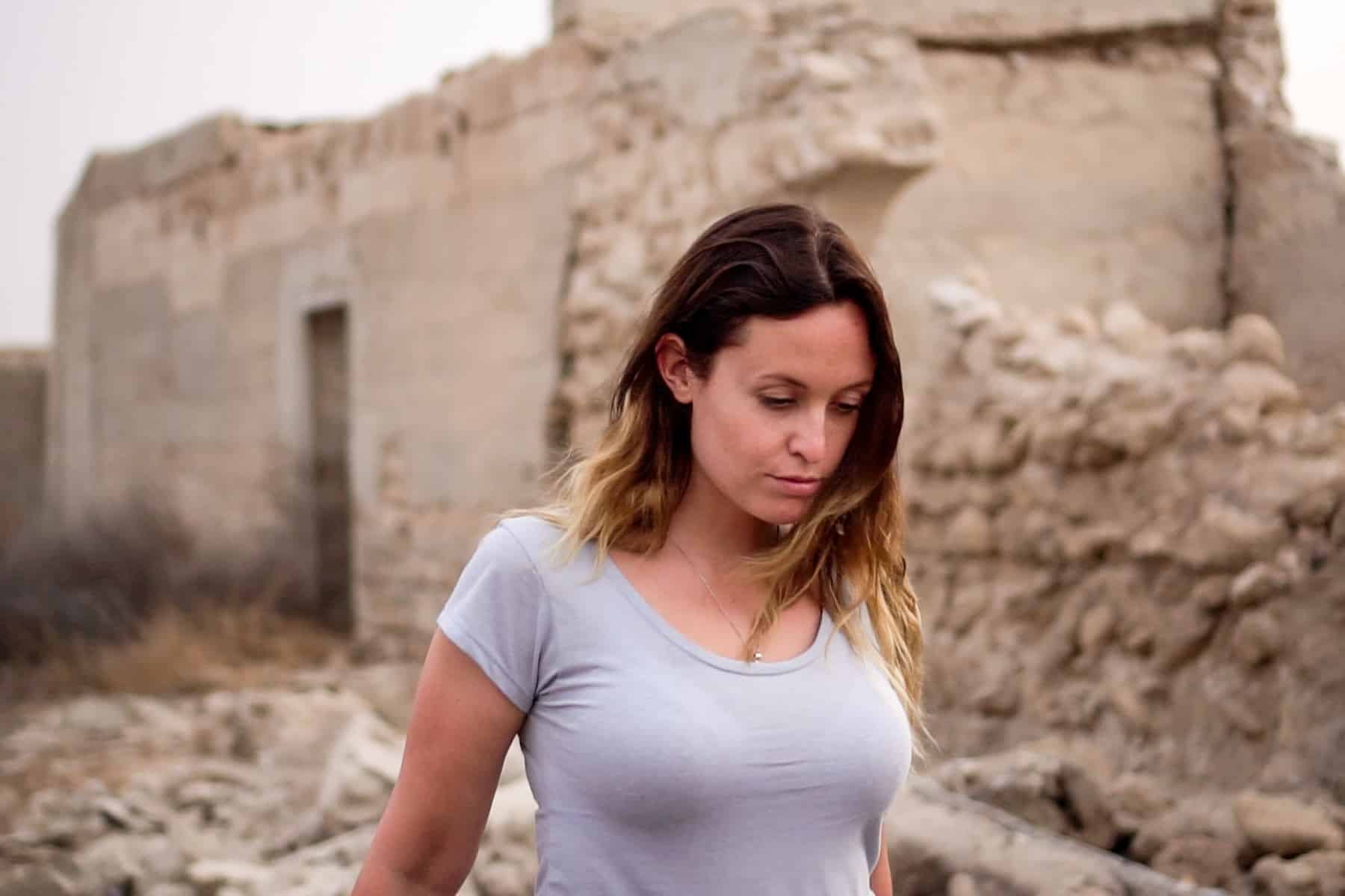 A woman in a blue t-shirt walks past an abandoned building and crumbling stone wall in the ghost town of Al Jazirat Al Hamra in Ras Al Khaimah 