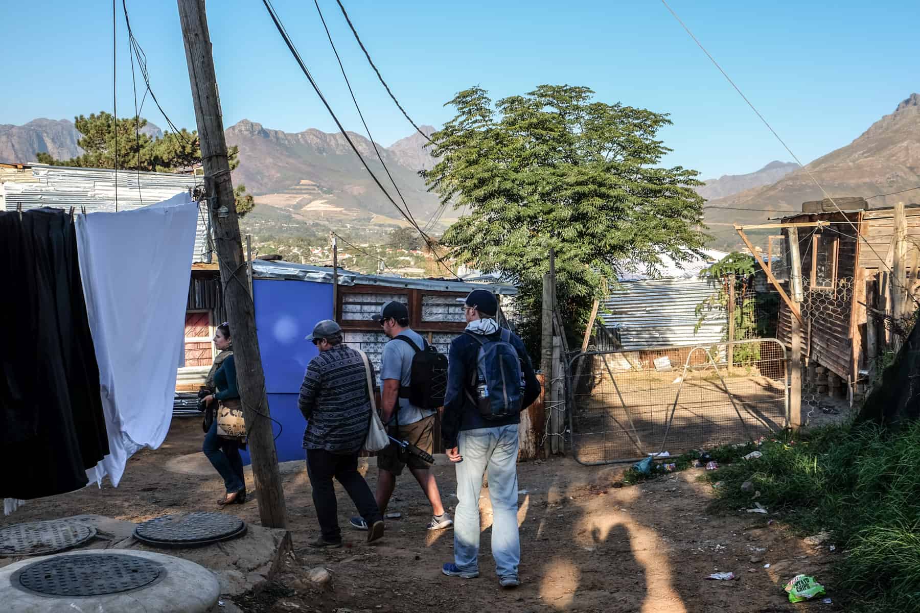 Four people walk through a dirt road on an enclosed street in a Township in South Africa, past hanging washing, low rise iron-clad housing and through rugged nature of trees, backed by mountains