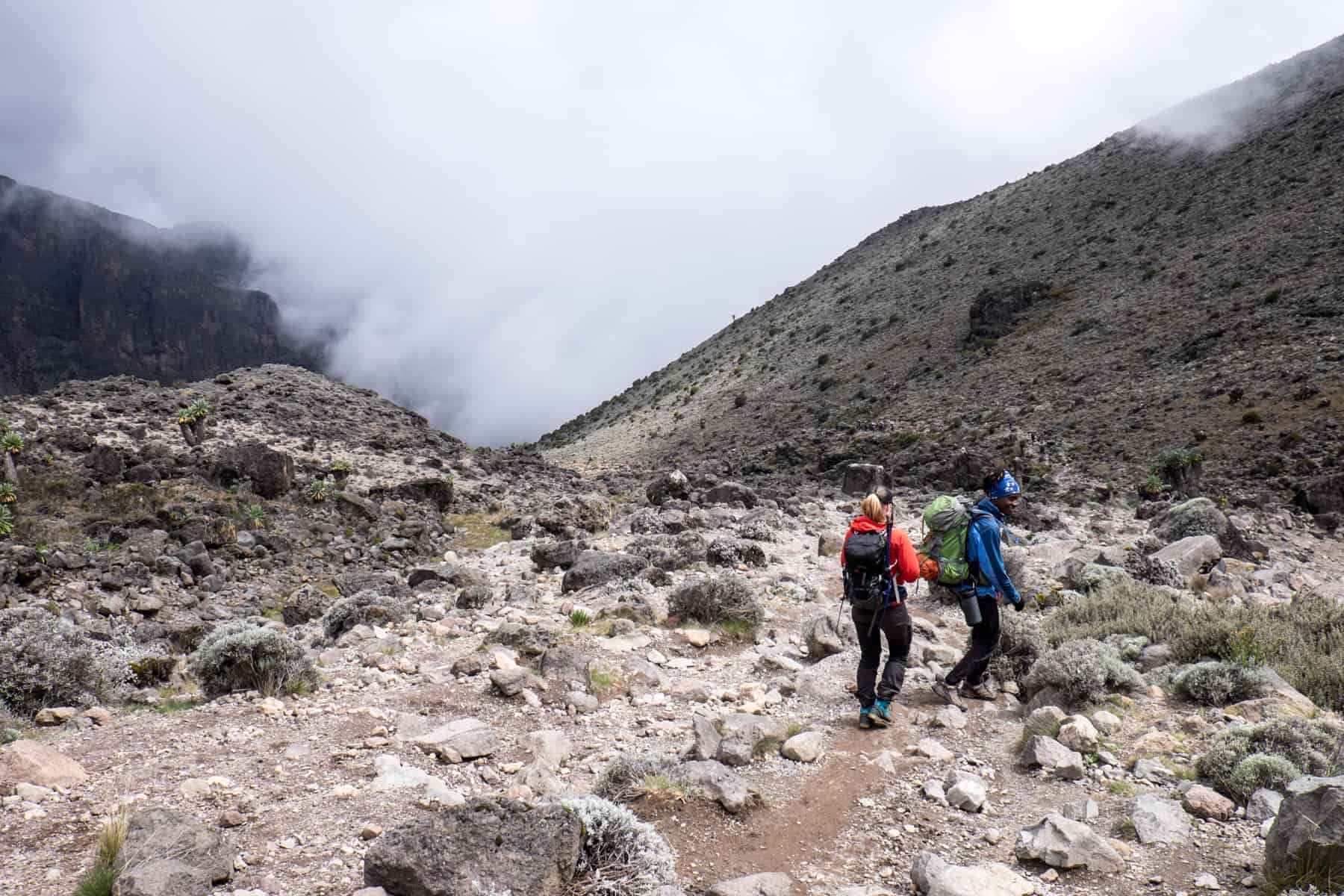 Two hikers in a rocky, mountainous plain with hovering cloud mist in the background.