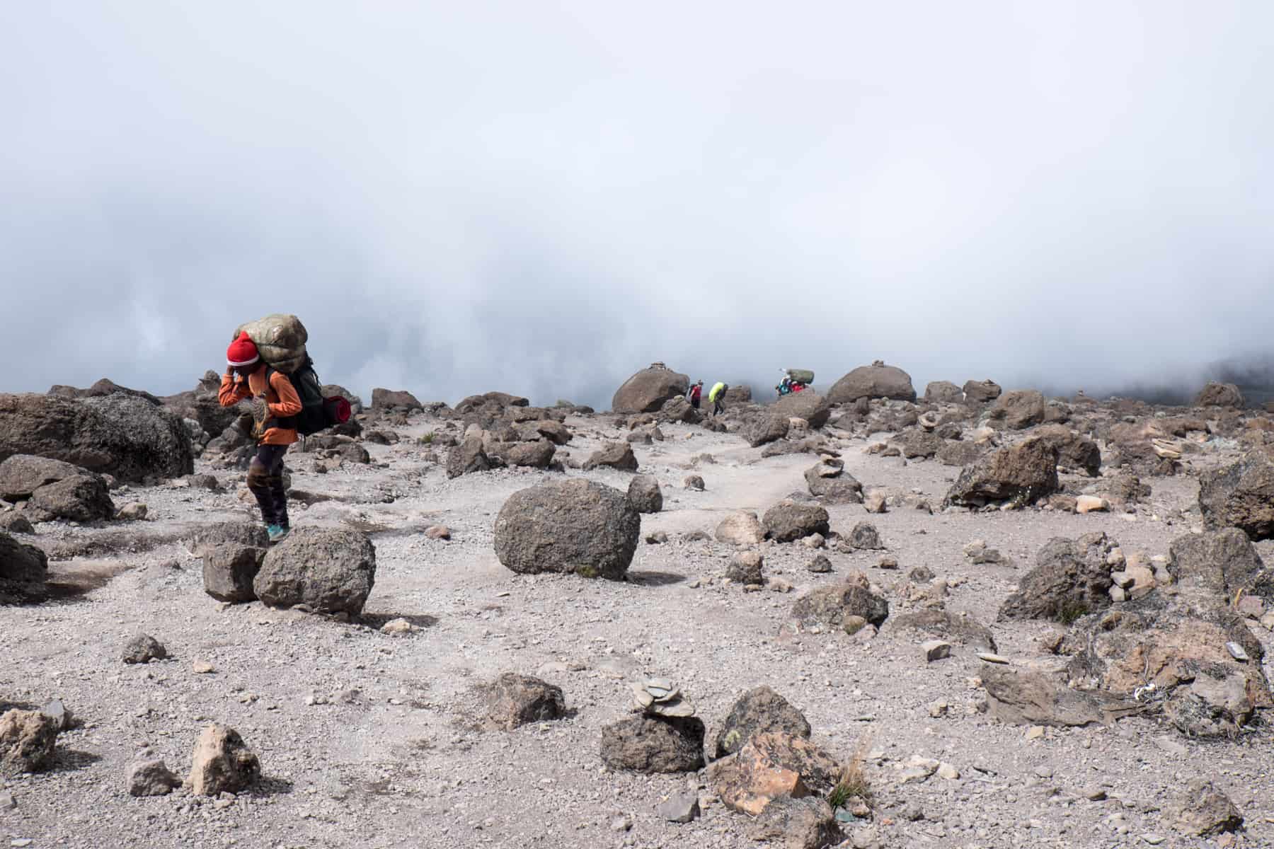 A Kilimanjaro porter wearing orange and carrying a large grey sack on his back walks through a barren grey-brown landscape strewn with large rocks. Mist rises behind him. 