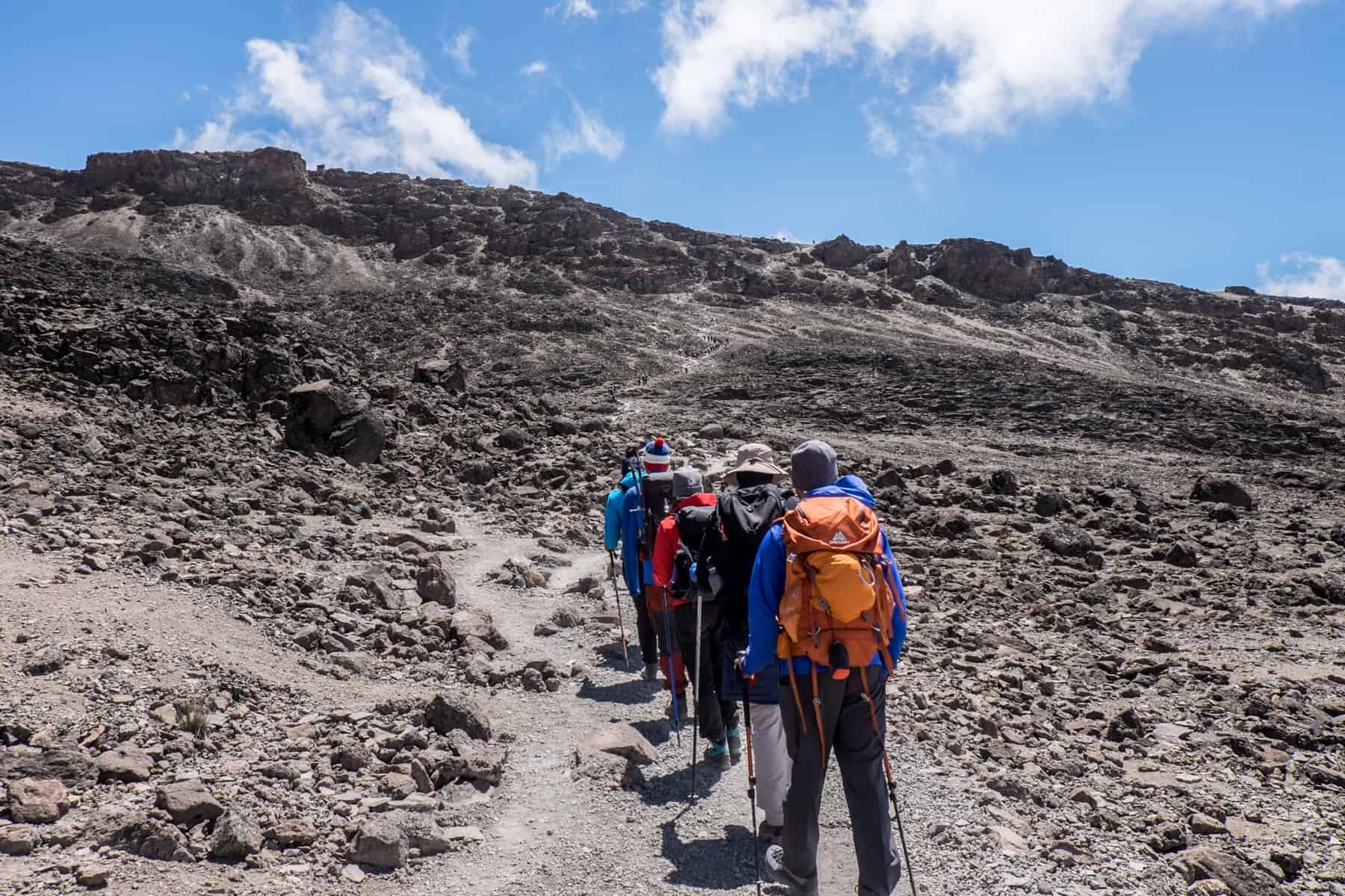 Kilimanjaro trekkers in colourful clothing walking on a flattened pathway on a steep, rocky slope. 