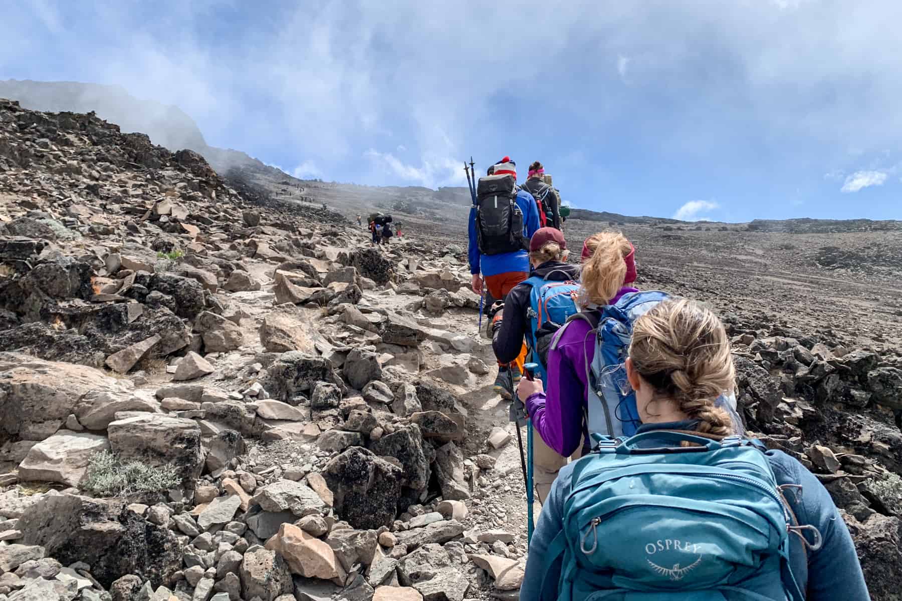 A line of five trekkers climbing Kilimanjaro's sandy coloured rocky wilderness towards a steep slope. Mist rises towards the blue sky. 