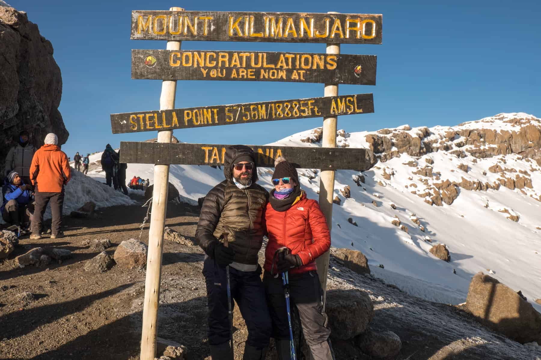 A man and woman, dressed in layers of hiking clothing, stand in front of the Mount Kilimanjaro Stella Point sign. Behind them is snow covered mountain slopes and other trekkers.