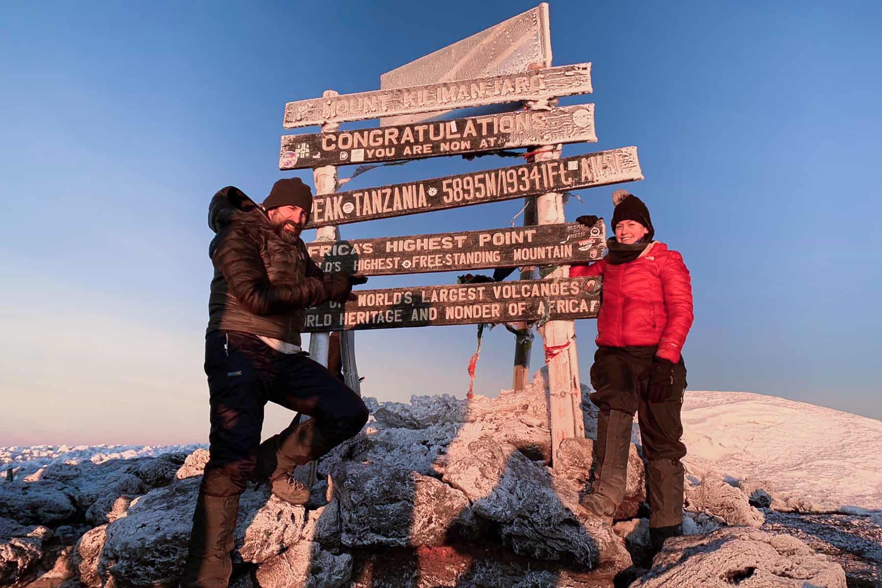 A man and woman stand in front of the mountain top, snow-covered wooden sign for Uhuru Peak after climbing Kilimanjaro.