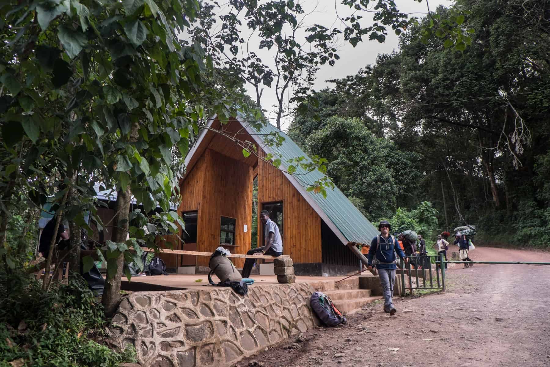 A man sitting on a bench looks towards the camera as another man, in a blue jacket, grey pants and a large hat smiles as he walks out of the wooden triangular building with a green roof - the end of the Kilimanjaro climb with the rainforest behind