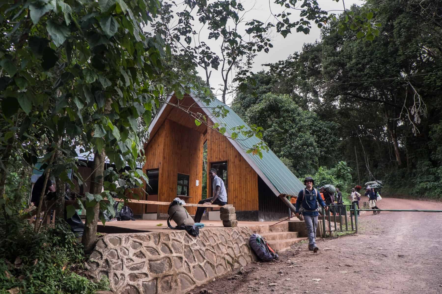 A man sitting on a bench looks towards the camera as another man, in a blue jacket, grey pants and a large hat smiles as he walks out of the wooden triangular building with a green roof, with the rainforest behind.