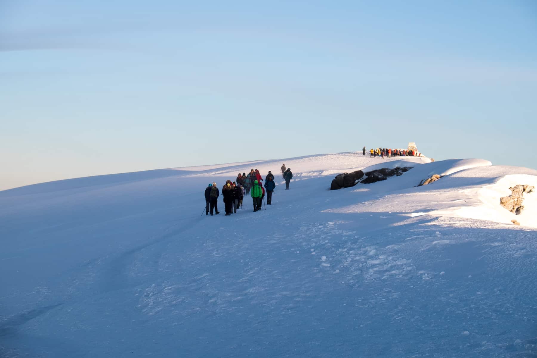 A small group of people walk towards the camera across a wide mass of pure white snow on the very top of Kilimanjaro mountain. In the background a group of people can be seen stationed around the sign board. 