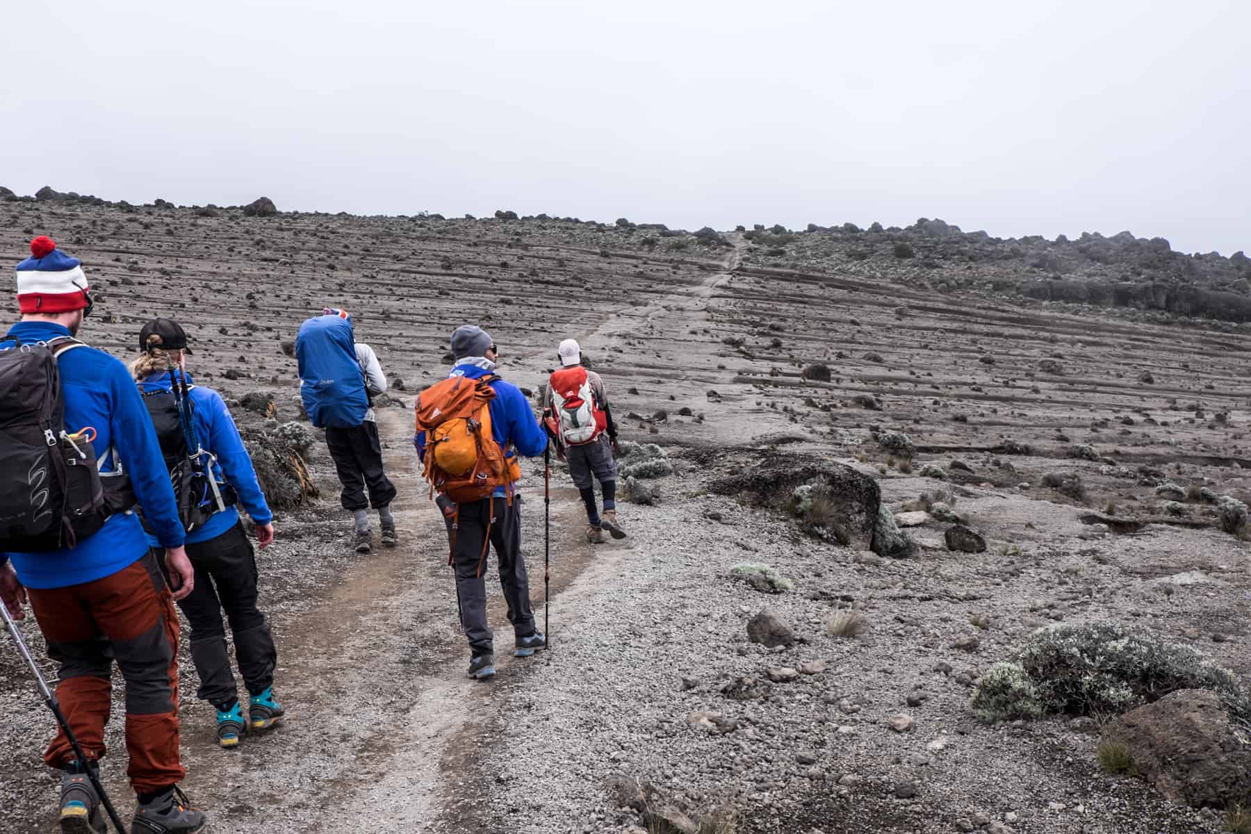Five people hiking Kilimanjaro, up a grey barren path with small dark rocks and towards a flat ridge