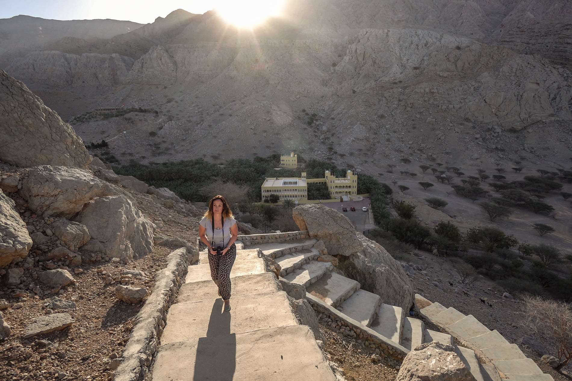 A woman walking up a stone staircase on a rocky hill with mountains behind.