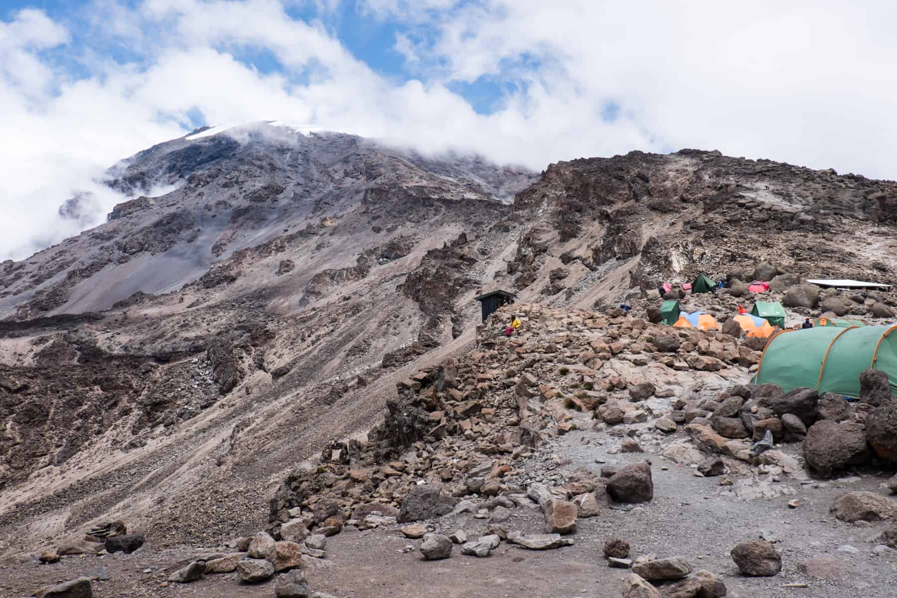 Kilimanjaro's snow capped, mist covered Kibo peak stands high on the left next to a bundle of colourful tents at Base Camp to the right.