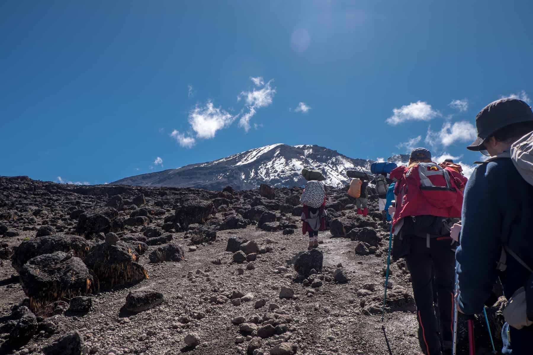 A line of trekkers walk on a dry, rocky pathway towards Mount Kilimanjaro that stands in the distance.