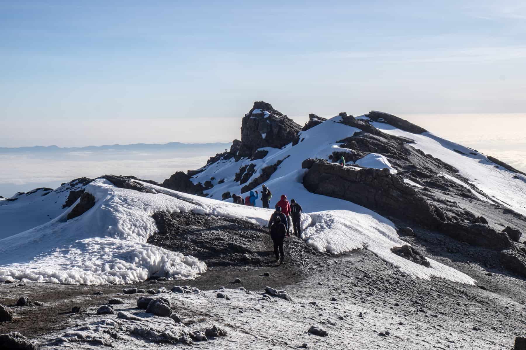 A small group of trekkers emerge from a path, onto the top of a rocky plateau of the Kilimanjaro Summit. The black mountain rocks are covered in patches of snow.