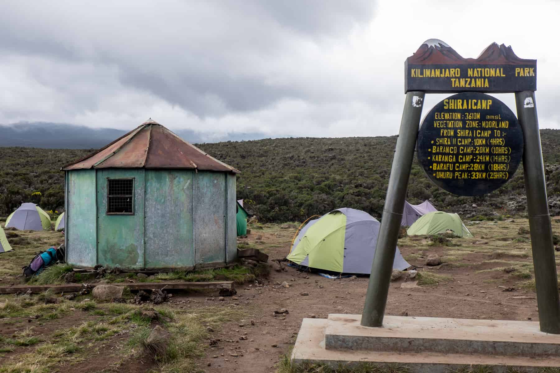 Brown Kilimanjaro Shira 1 Camp Sign next to a mint green octagonal building with iron roof. A small camp can be seen behind them. 