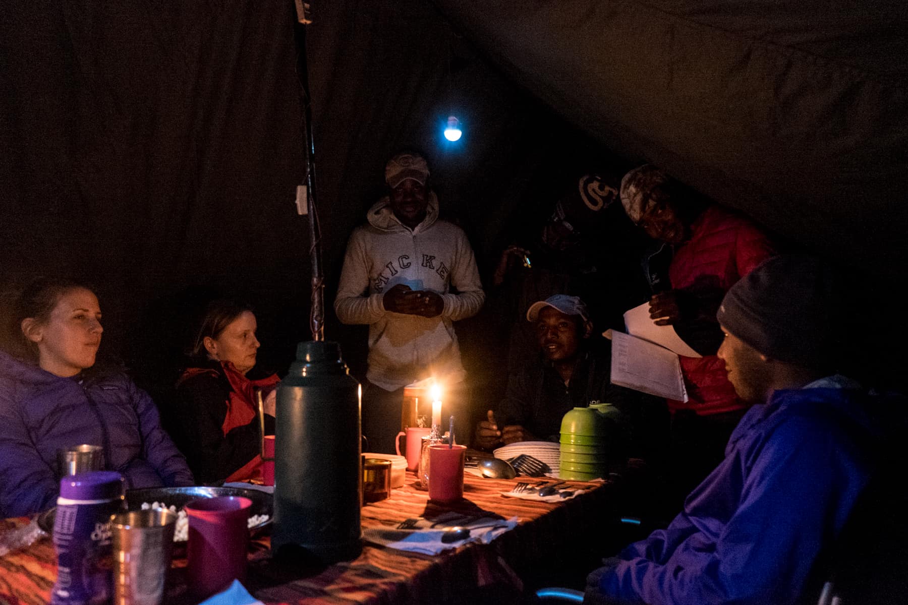 A group of Kilimanjaro trekkers sit round a table in a tent at night, while four guides give a briefing