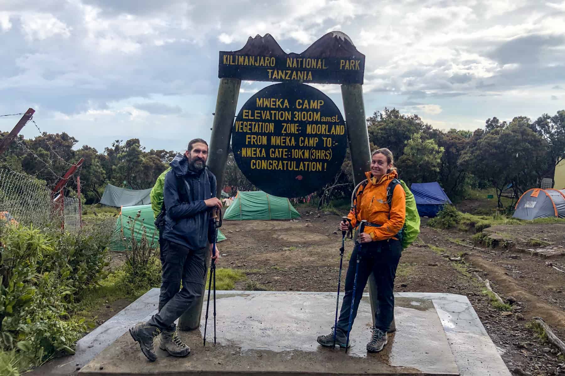 A man and woman, looking extremely tired and holding walking poles, stand in front of the Kilimanjaro Mweka Camp wooden sign. A scattering of tents can be seen in the background. 