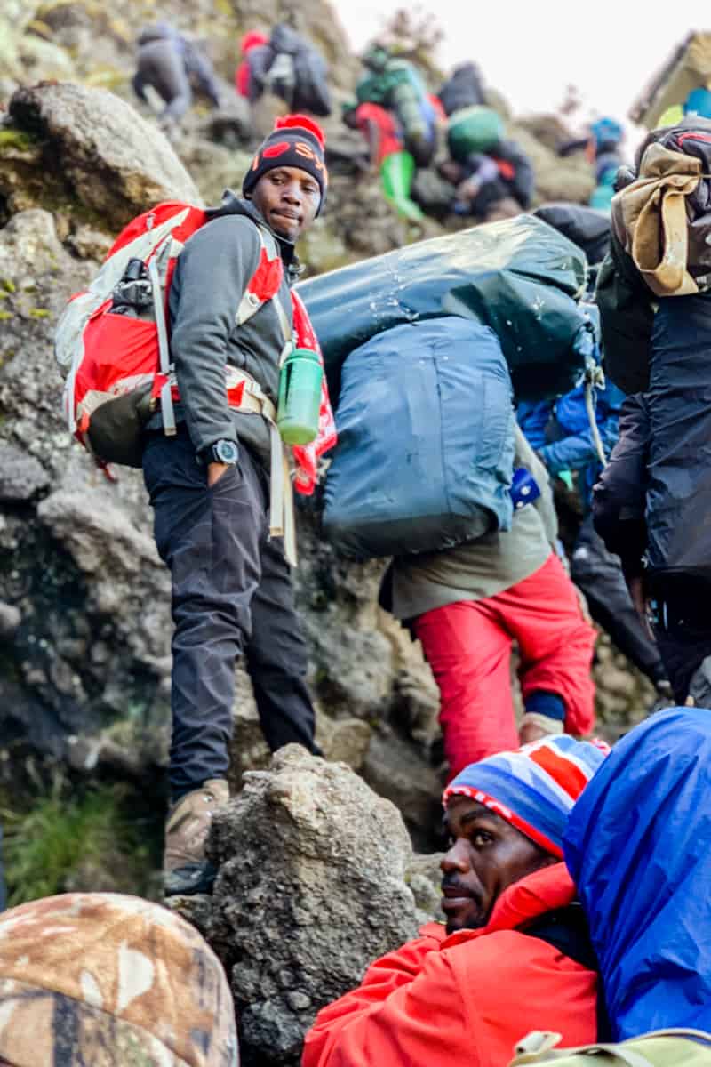 A guide dressed in grey with a red backpack looks on seriously as trekkers climbs past him on the high Barranco Wall on the Kilimanjaro trek
