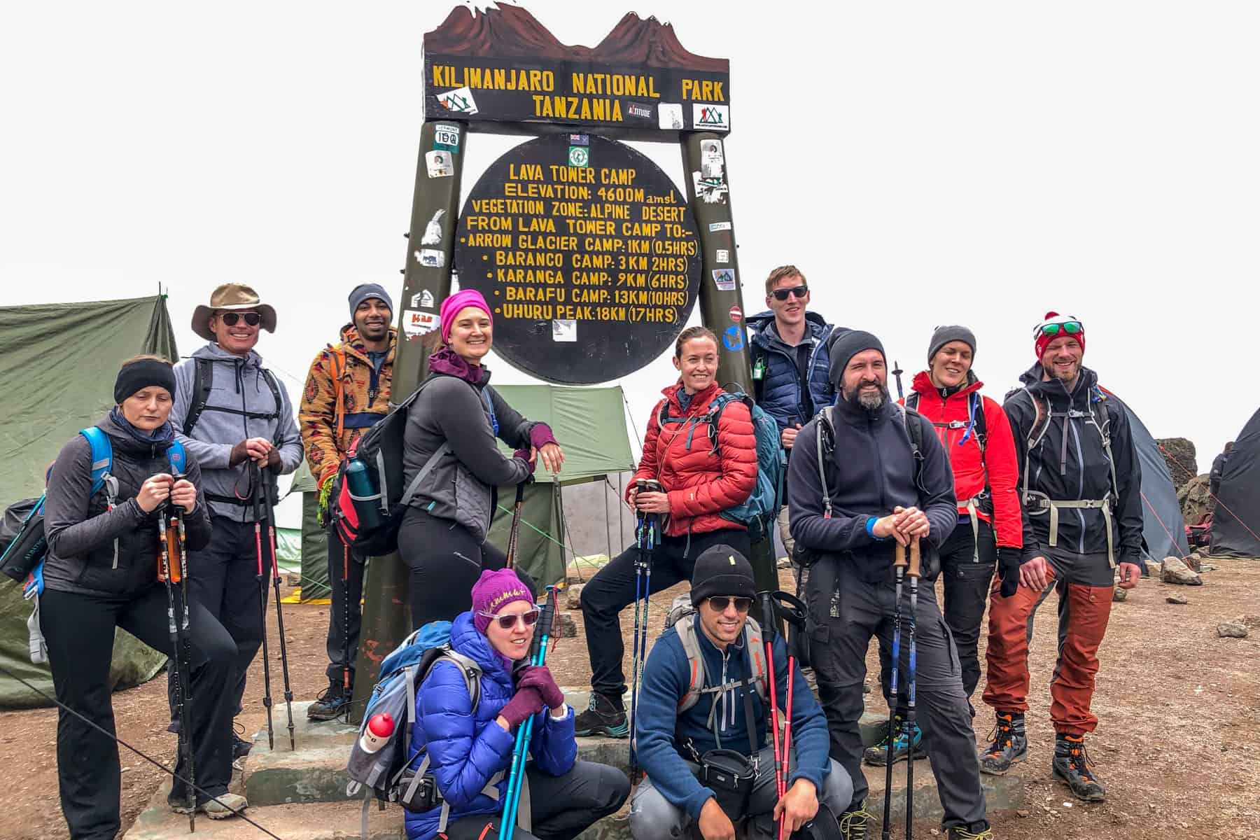 A Kilimanjaro trek group, looking tired, at the Lava Tower Sign with green camp tents set up behind them.