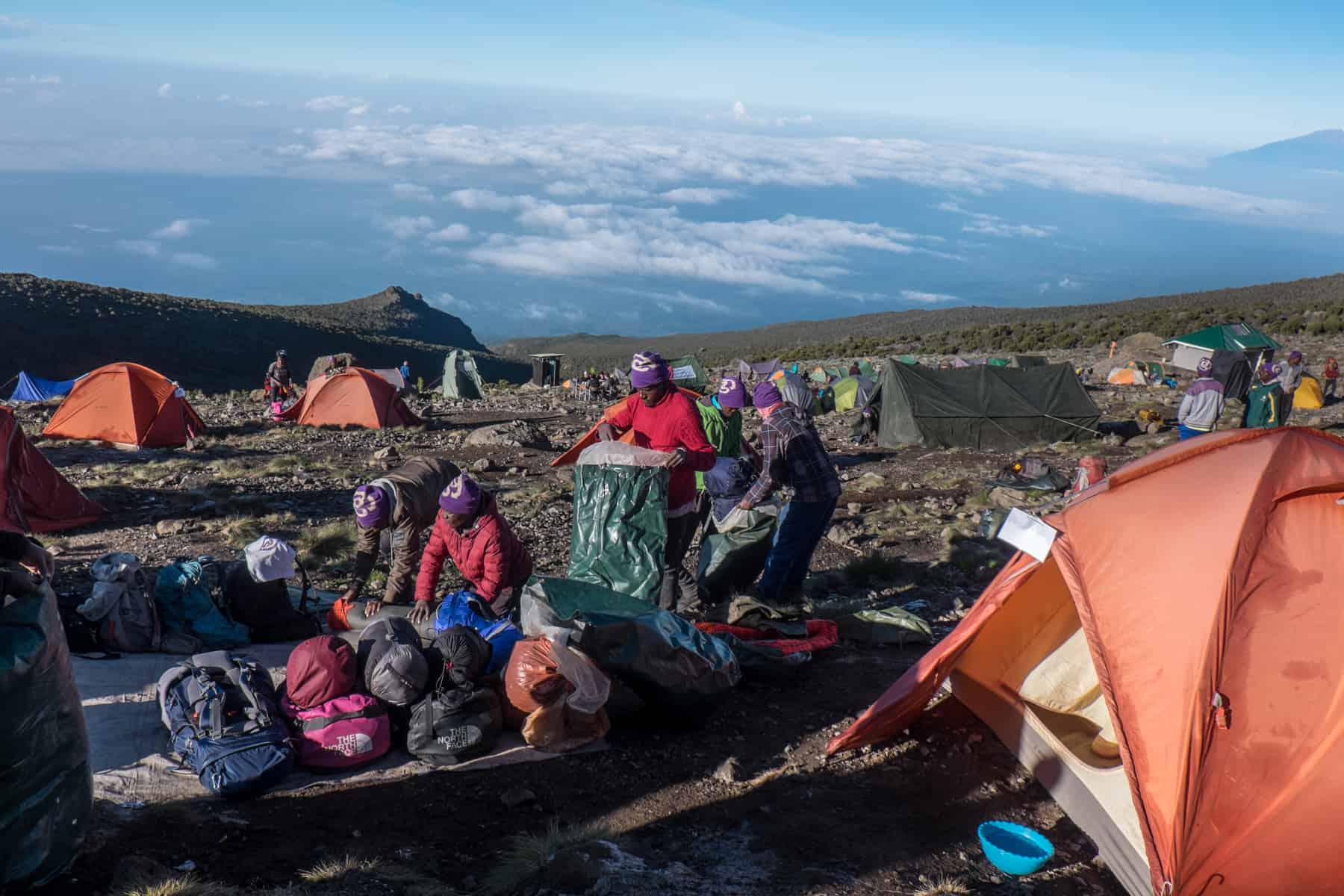 Porters in purple hats at a Kilimanjaro camp with orange and green tents, packing away equipment. 