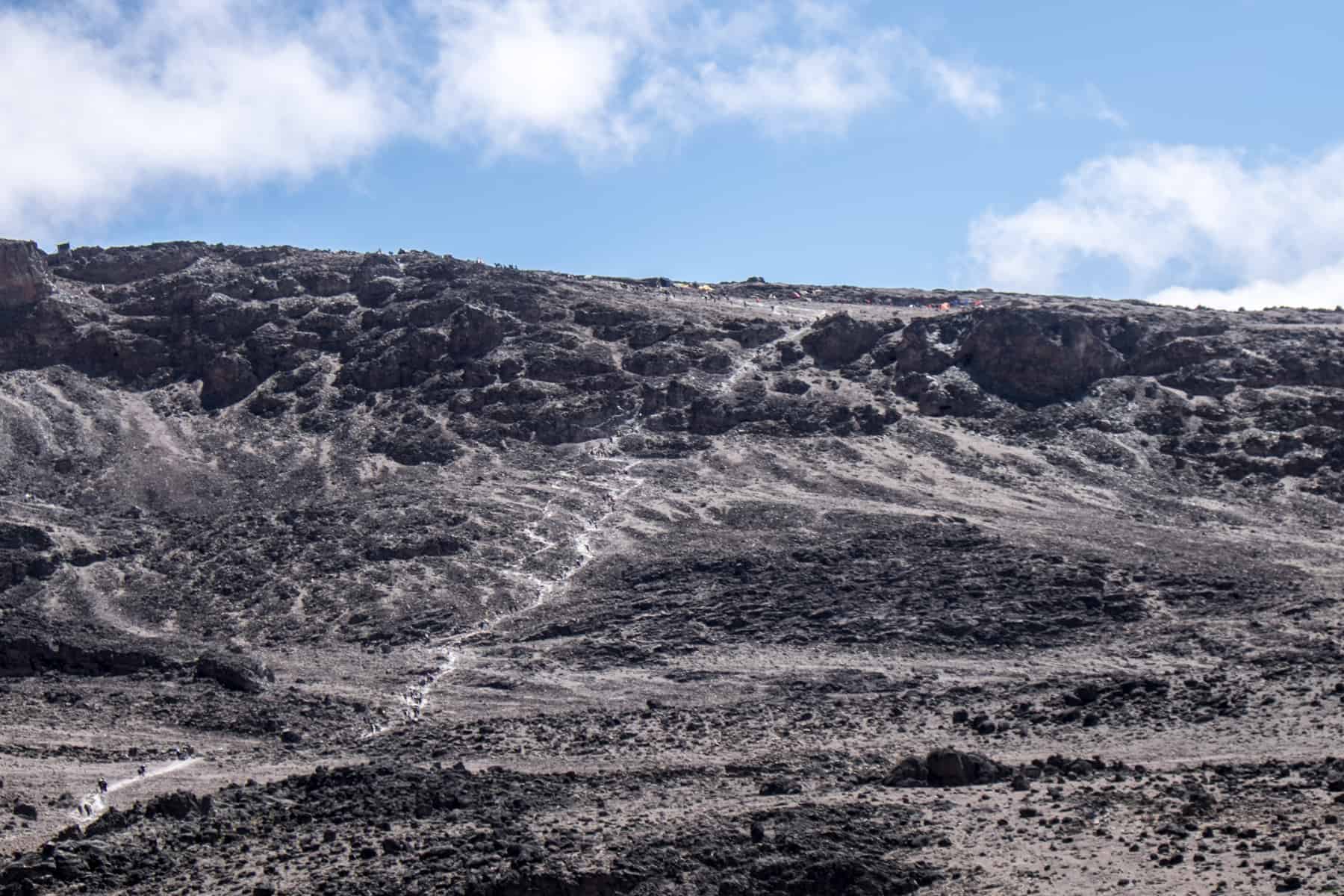 A tall, black volcanic mastif looms under a blue sky. White walking trails, part of the Kilimanjaro trek, stretch from top to bottom 