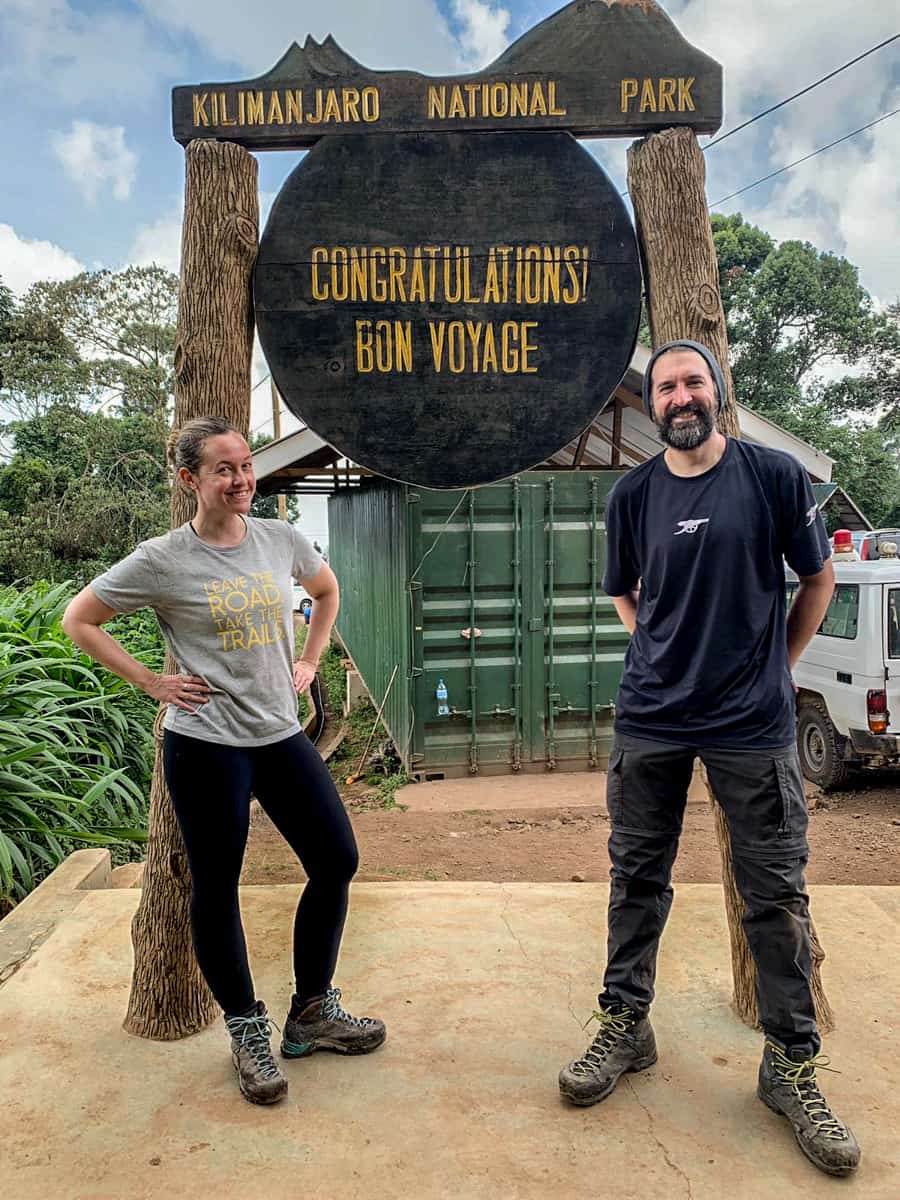 A woman in grey and man in black, standing in front of the Kilimanjaro National Park 'Congratulations! Bon Voyage' signage at the end of the trek.