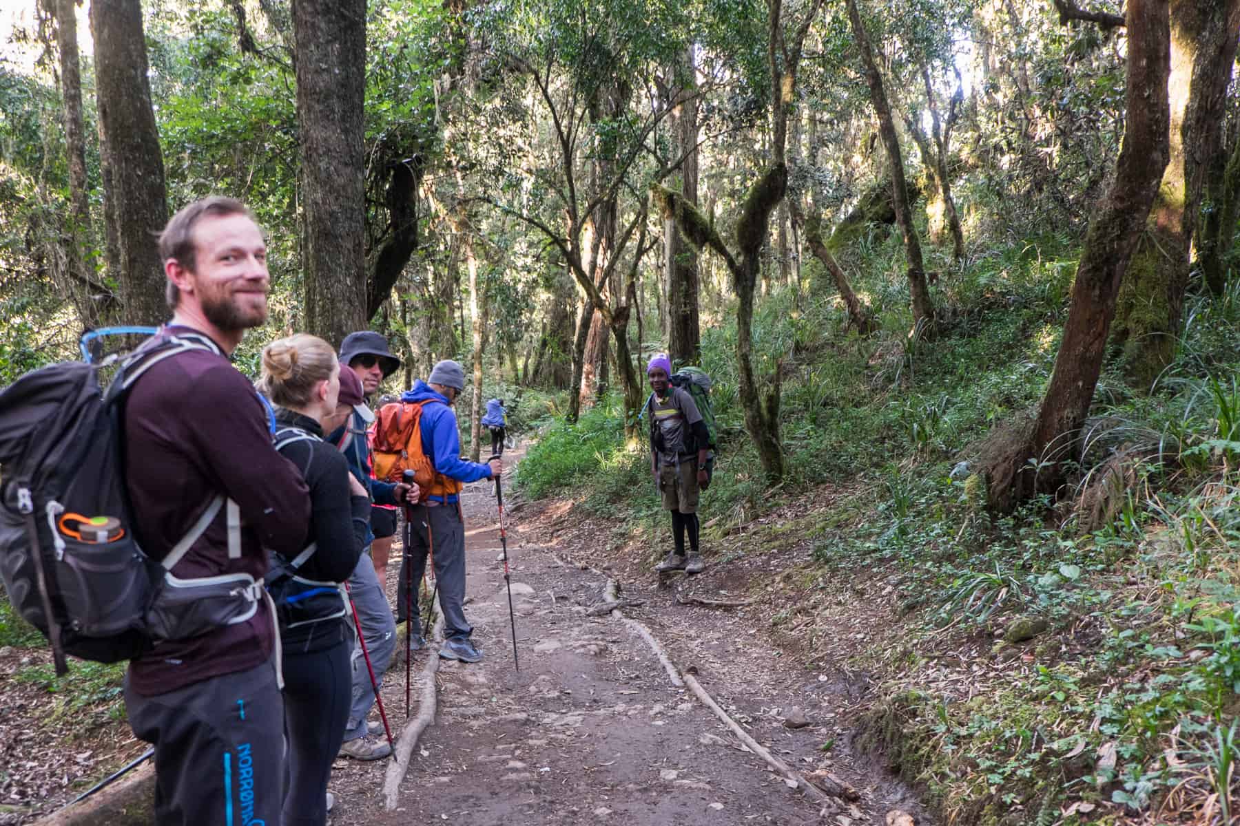 Five trekkers stand the left of a muddy trail path, and a guide, wearing a purple hat and carrying a large bag stands to the right, in the Rainforest zone of Kilimanjaro.