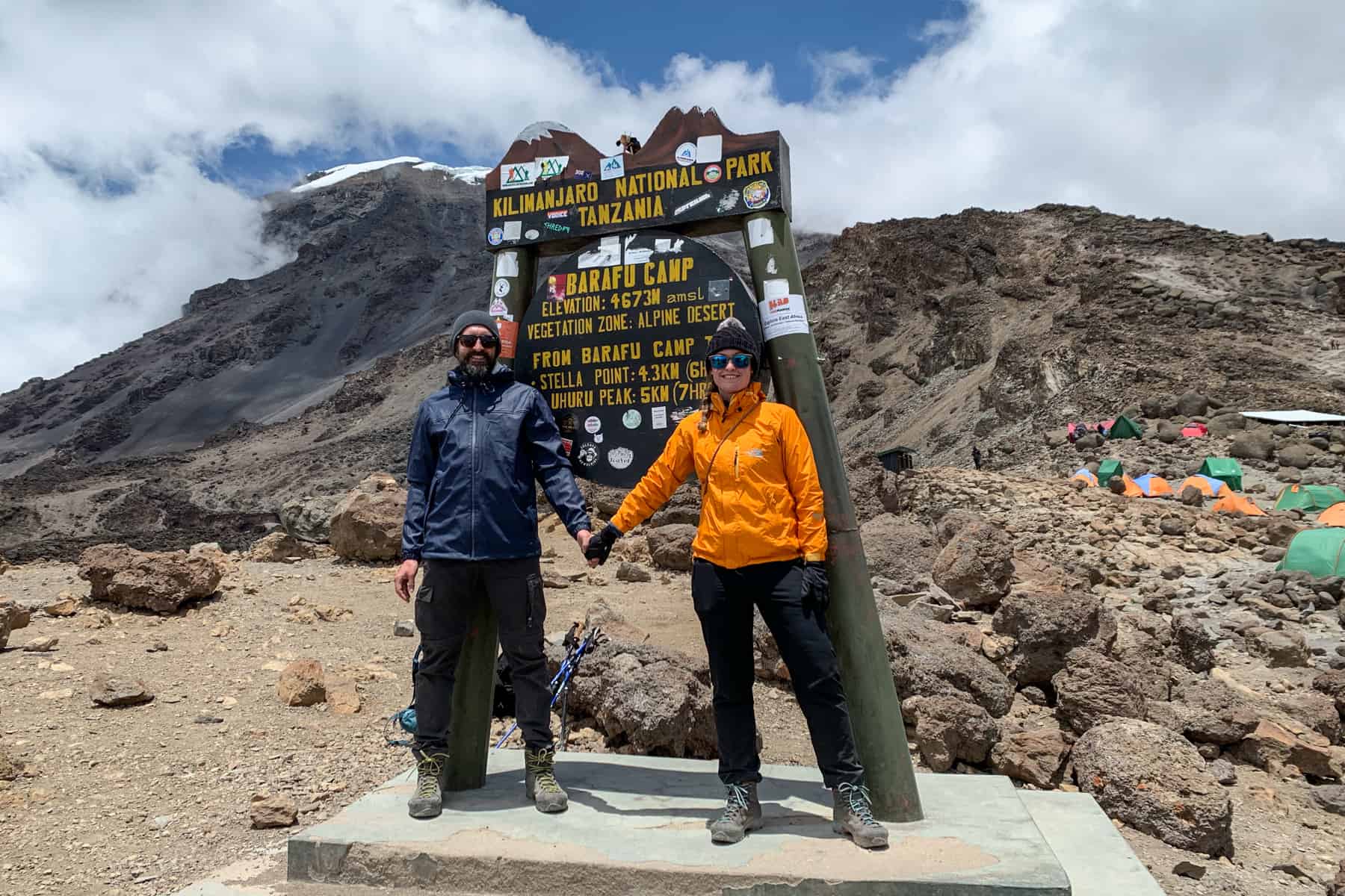 A man in dark trekking clothing, and a woman in an orange jacket standing at the Barafu Camp sign on the Kilimanjaro trek to a backdrop of snow capped mountains. 