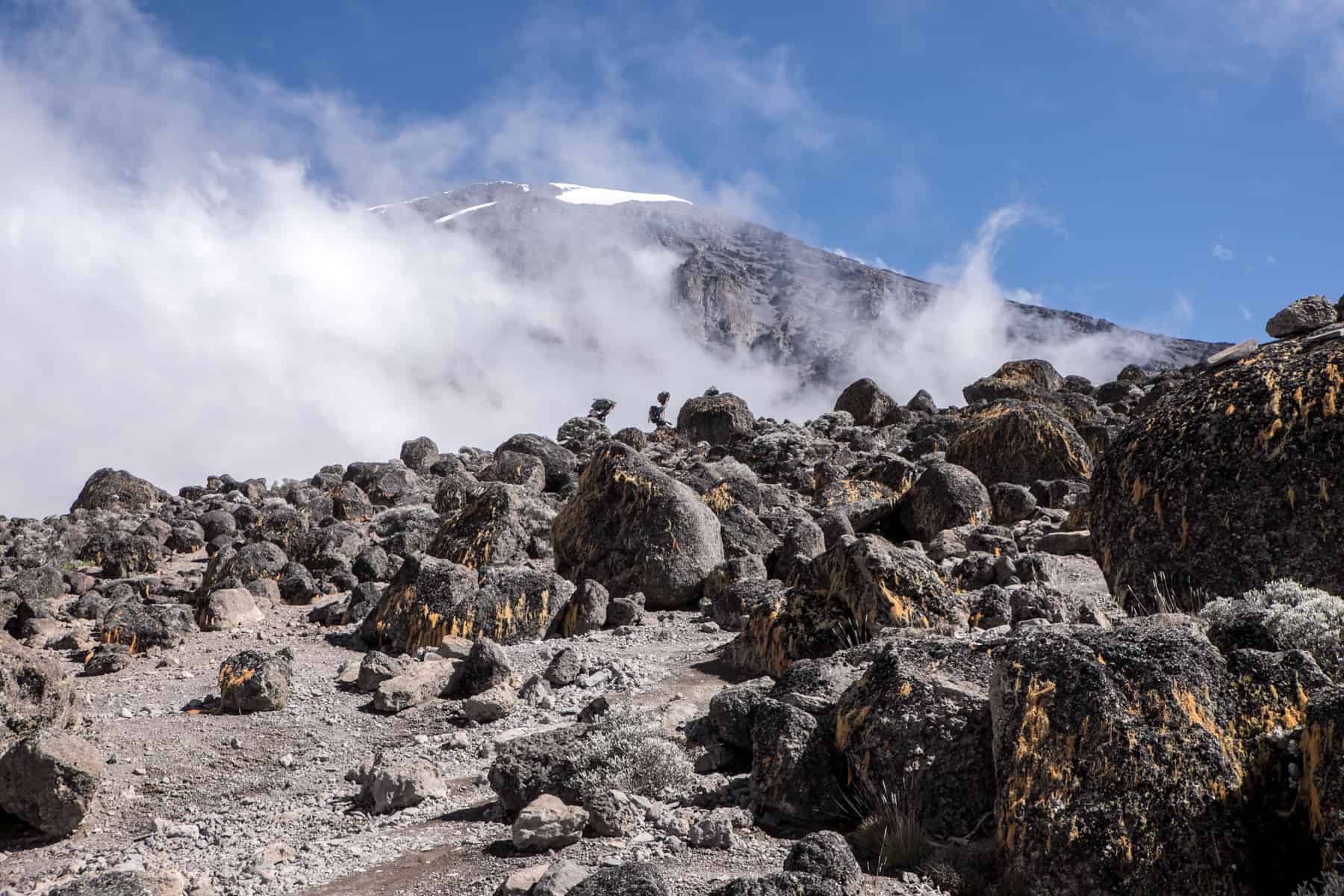 Porters, carry large bags on their heads, walk over the rocky volcanic landscape of Mt Kilimanjaro in front or rising mist and the mountain peak.