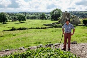 Farmer, Joe O'Connor in a blue shirt and deep-red trousers leans on a stick on a gravel path. Behind him is a vast stretch of green land scattered with sheep and a mound of grey rock ruins of the lost city of Jerpoint Park in Kilkenny, Ireland.