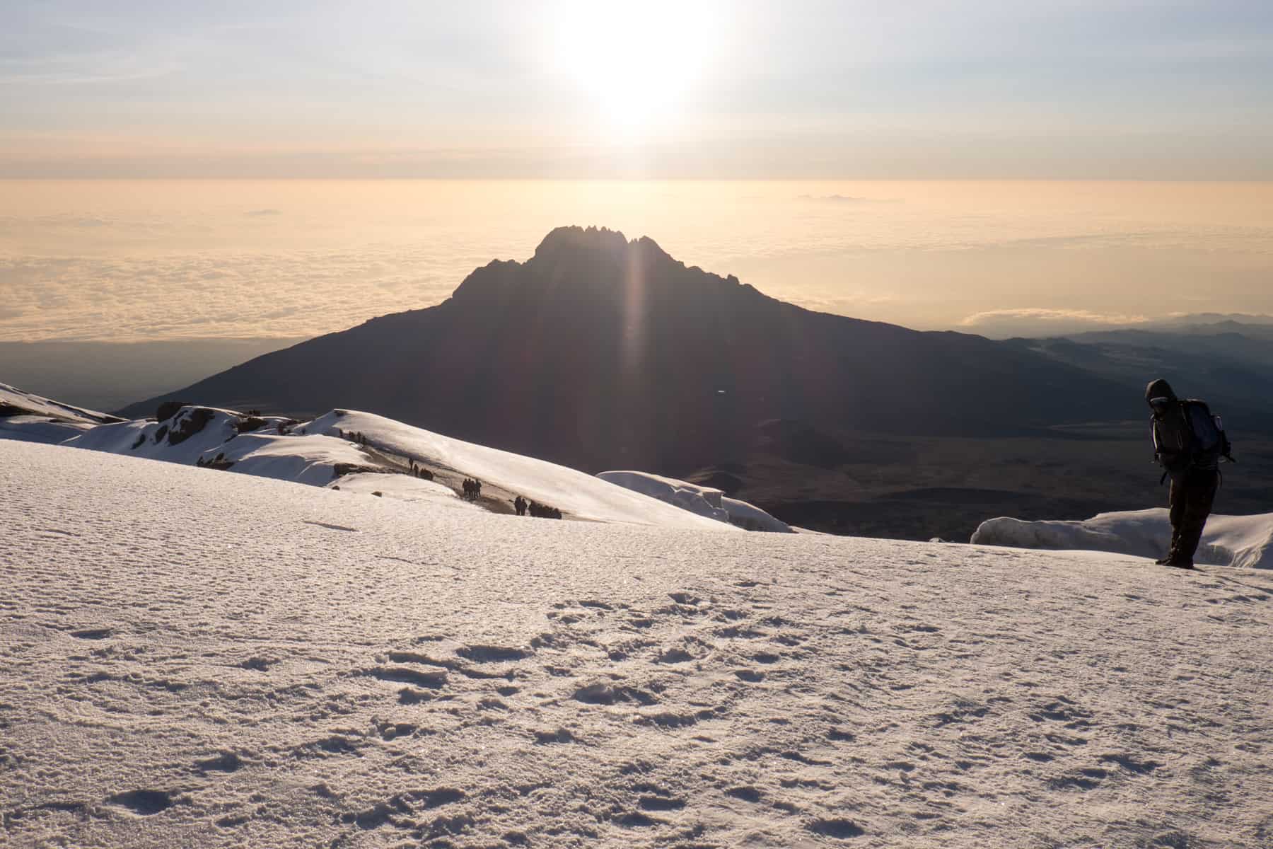 A man dressed in winter gear stands on the Summit of Kilimanjaro mountain on thick, white snow as the light yellow sun rises above a smaller peak.