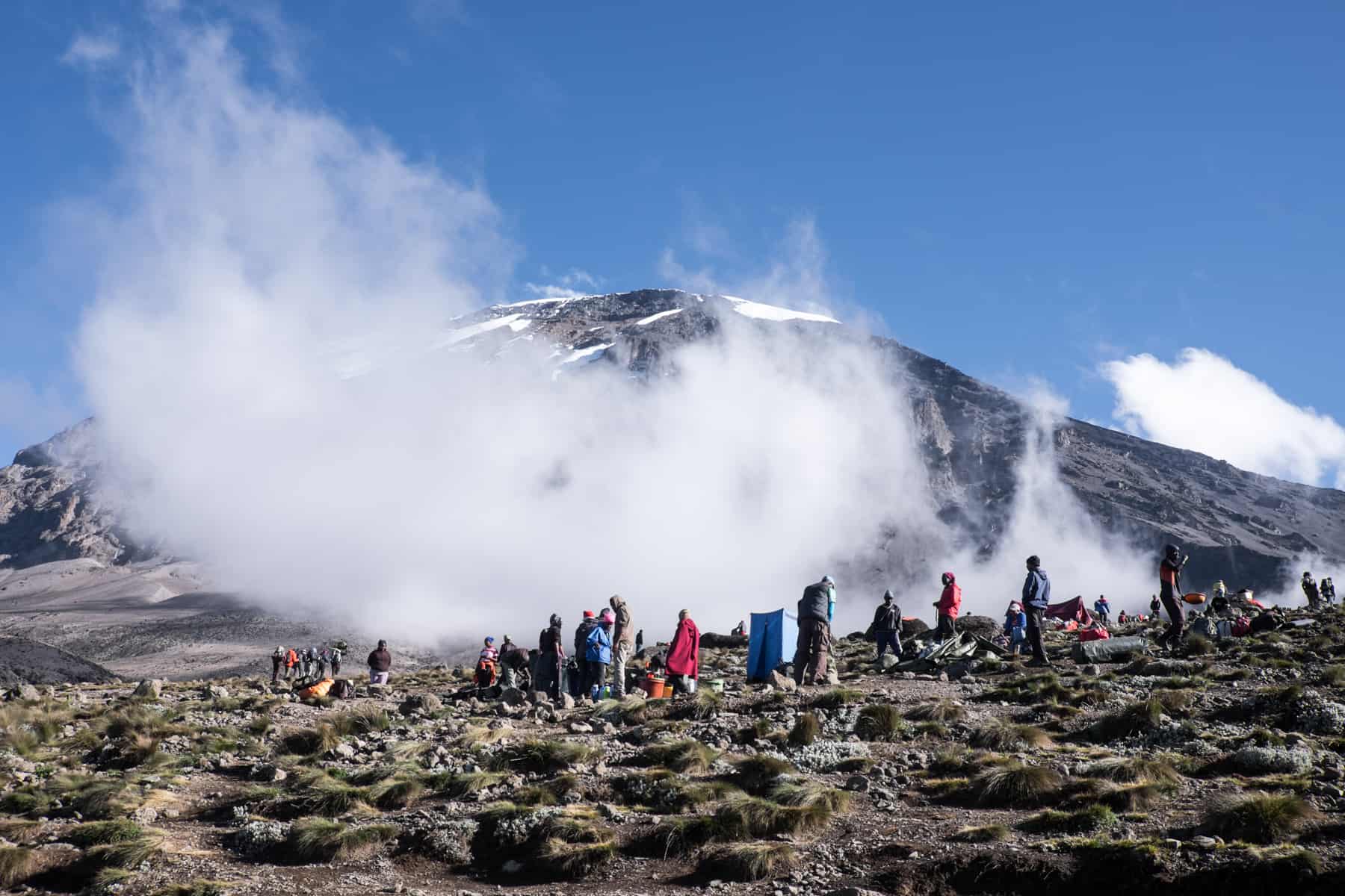 A group of Sherpa's packing away tents on a campsite in front of the looming Mount Kilimanjaro, engulfed in mist.