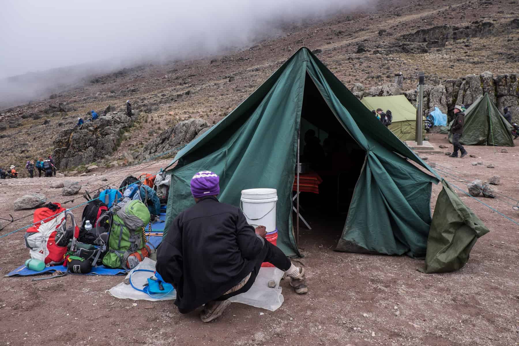 A porter on the Kilimanjaro trek sitting outside a green tent and filling water bottled from a white water barrel