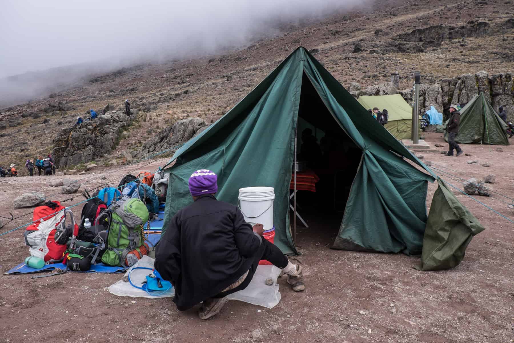 A porter on the Kilimanjaro trek sitting outside a green tent and filling water bottled from a white water barrel.