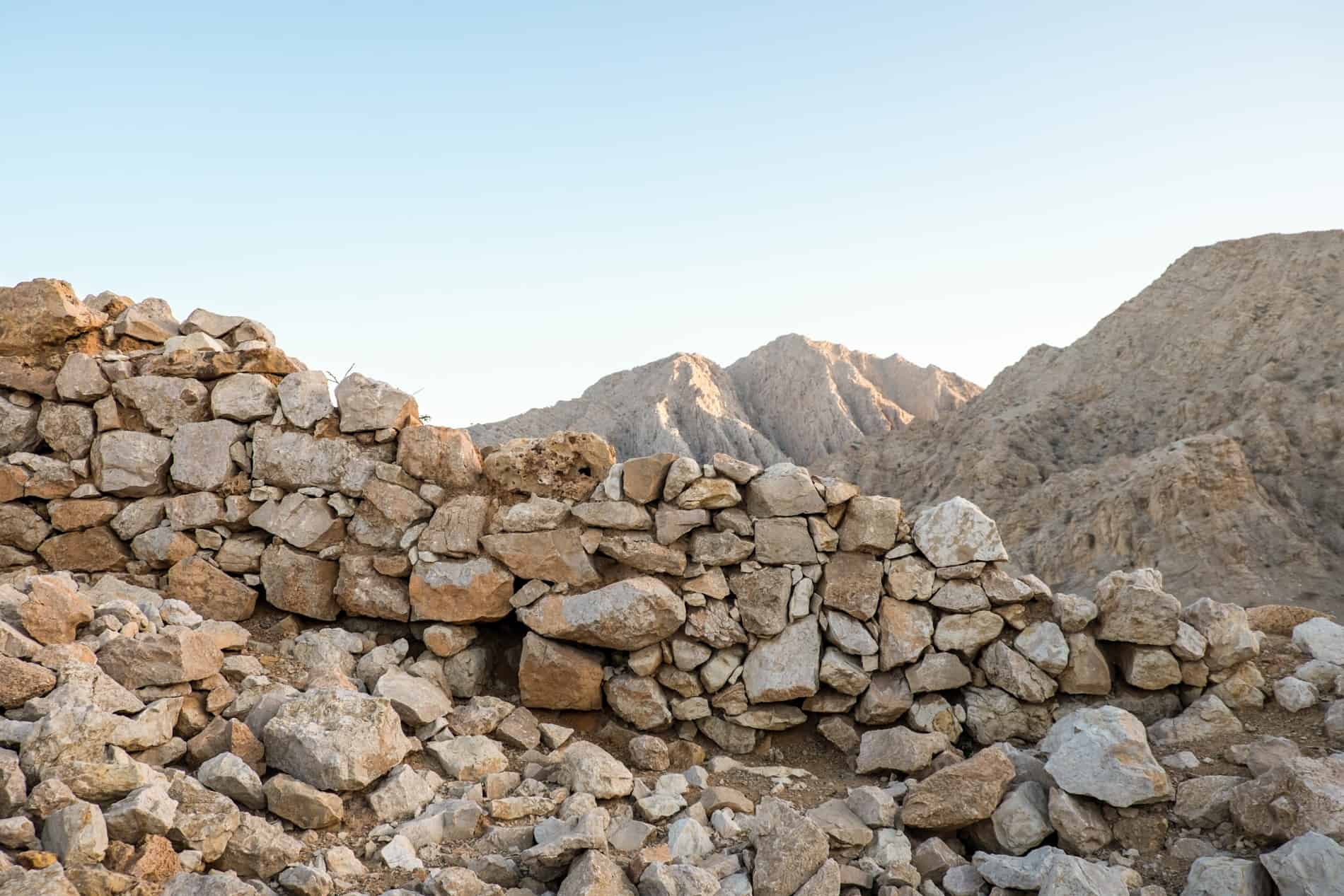 A sandy coloured rock stone wall with the Jebel Jais mountains in the background. 