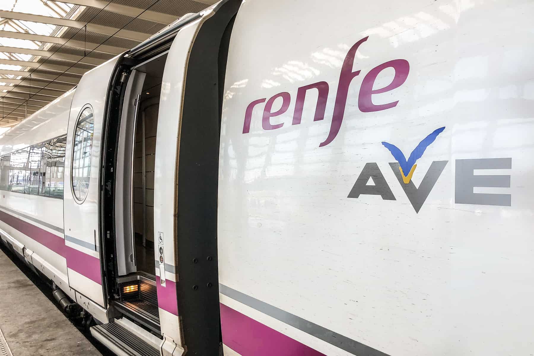 Close up of a white with pink stripe Renfe AVE train in Spain, waiting at a platform with open doors. 