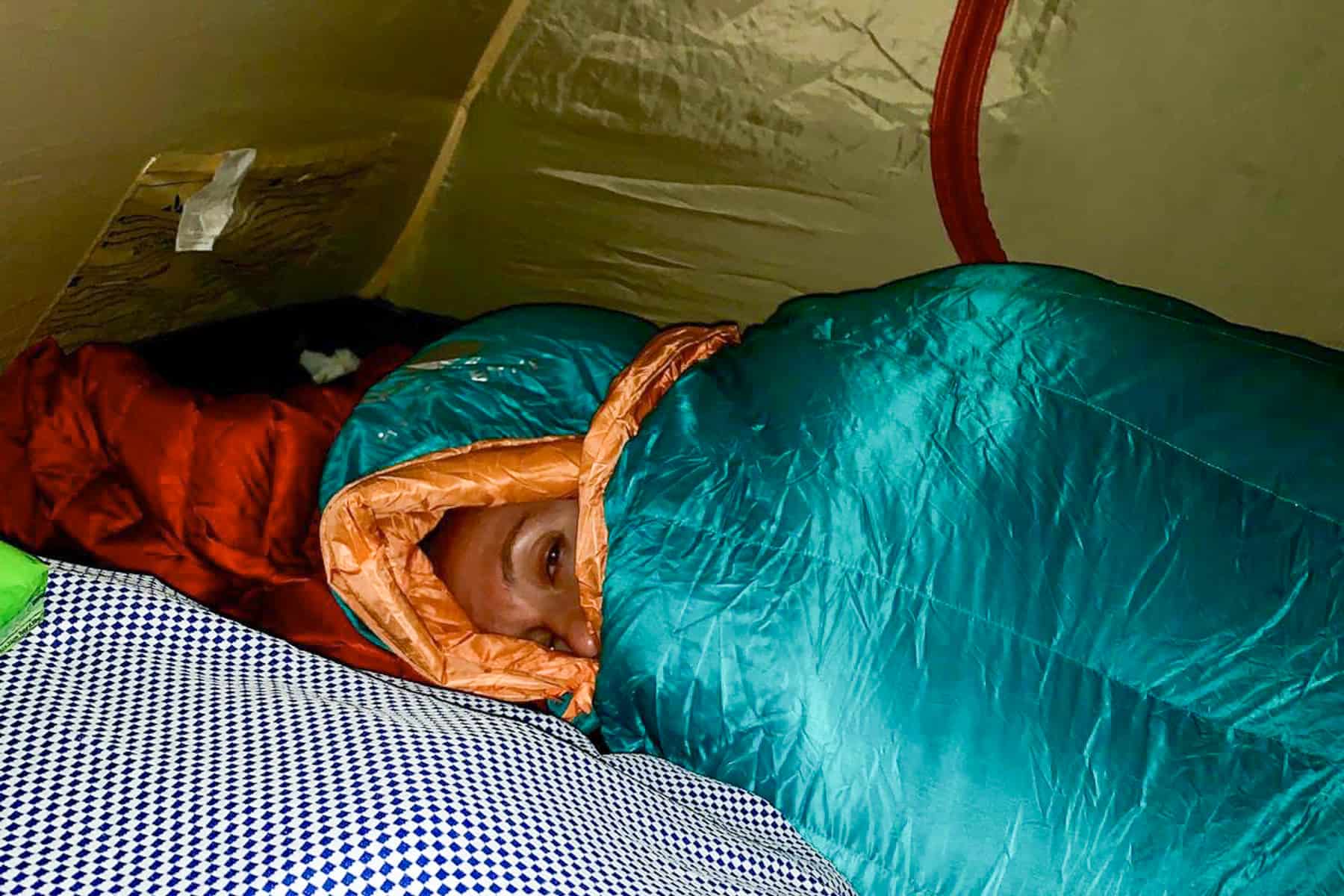 A women lies snug in a mint green and orange sleeping bag in a small tent during a Kilimanjaro trek