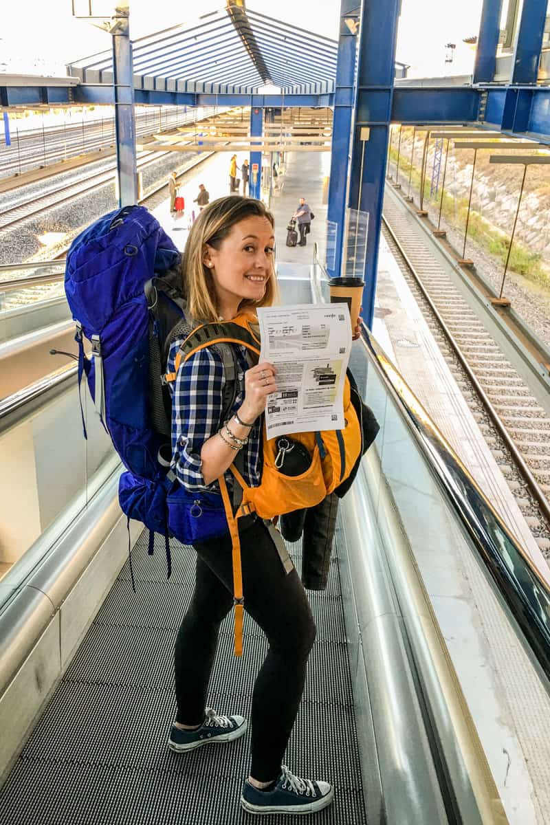 A woman with a blue backpack and yellow front pack, holding a printed ticket and a cup of coffee, on an escalator leading to a train platform. 