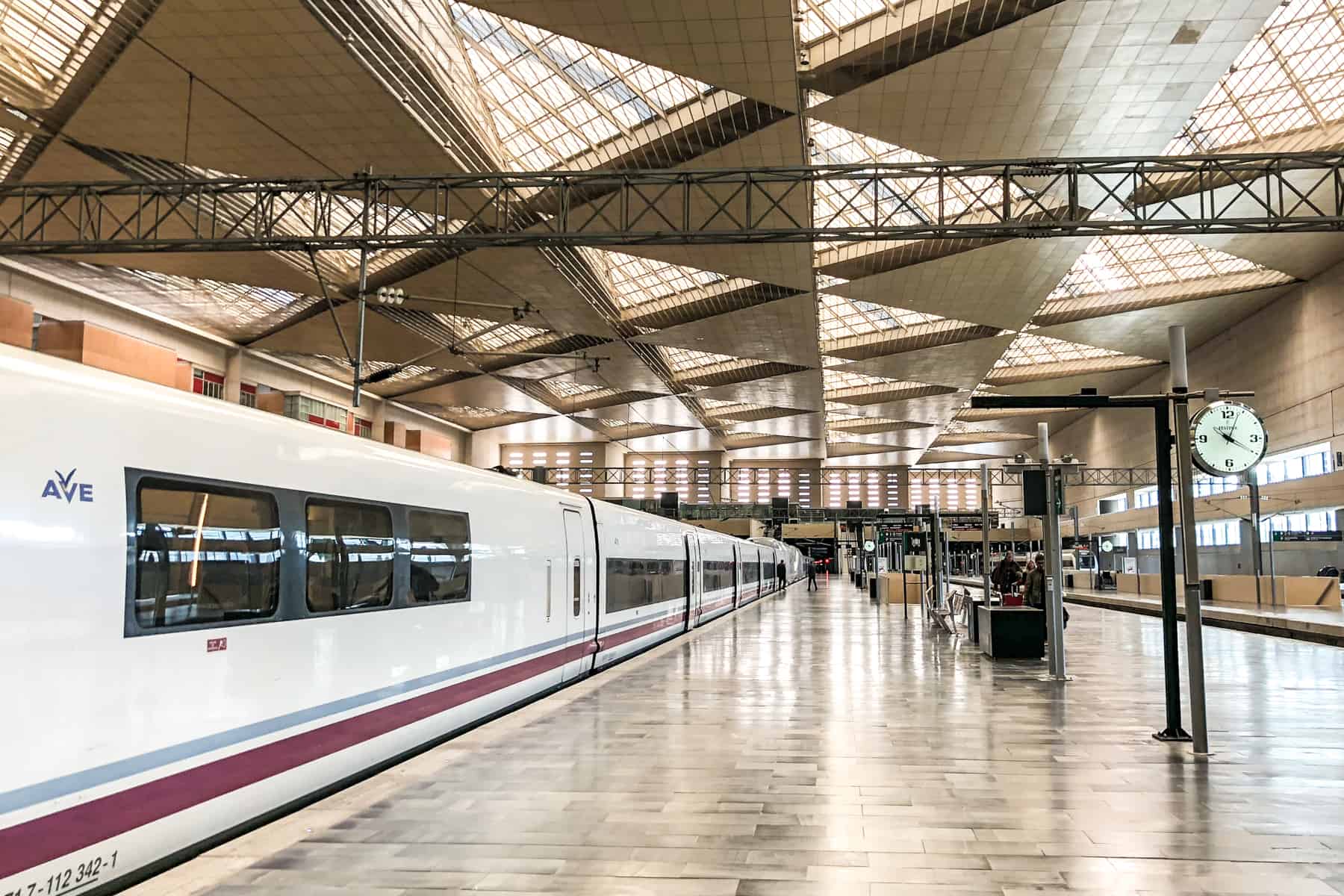 A long, white train with maroon and blue stripes and the letters 'Ave' stands at a modern train platform in Spain.