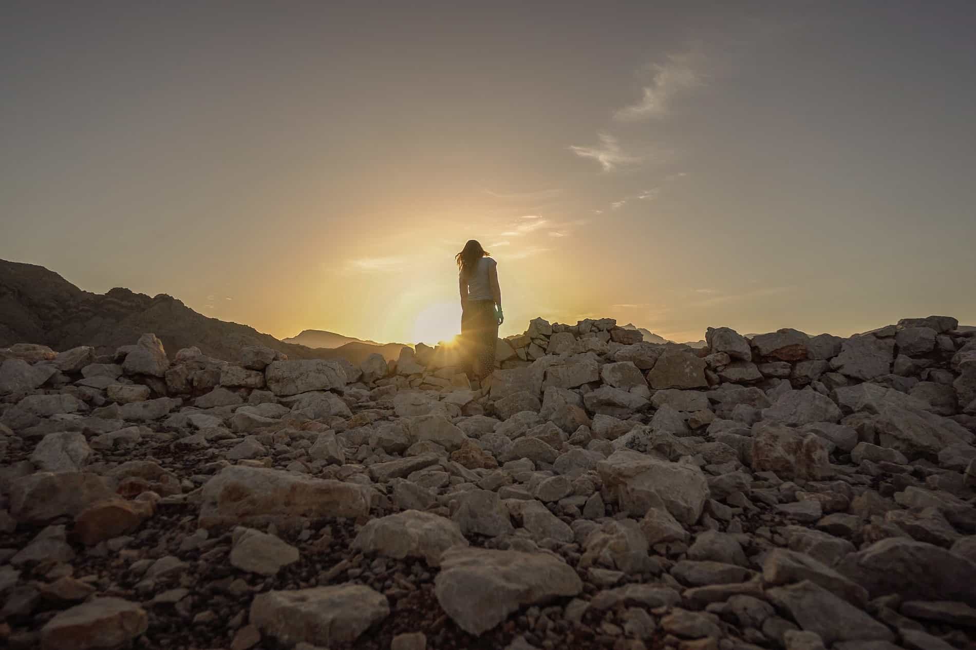 A woman stands upon a mound of rocks looking towards the sun as it rises above the landscape. 