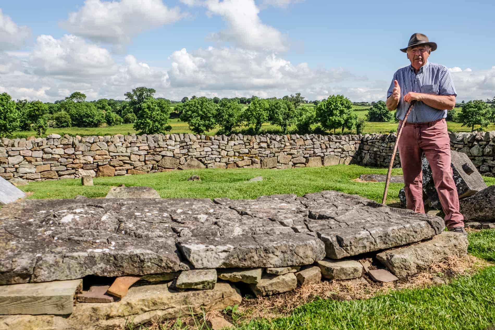 A man in red chinos, checkered shirt and hat stands with a stick next to the stone tomb of St. Nicholas in Jerpoint Park.