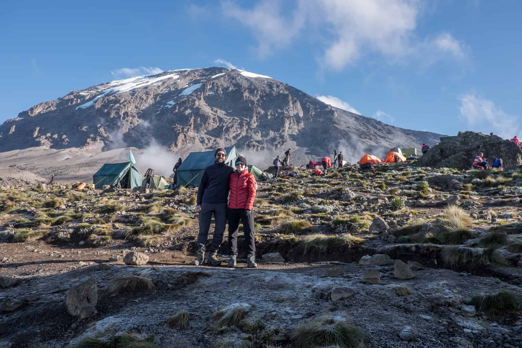 A man and woman stand together in front of a camp of green and orange tents, with the snow capped Mount Kilimanjaro behind.