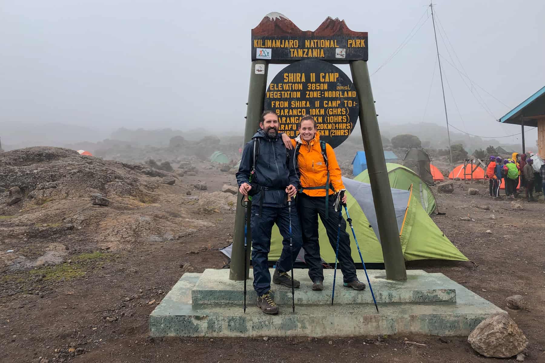 A man in blue and a woman in orange standing at the brown "Shira II Camp" Kilimanjaro sign with tents and people in the background shrouded in mist.