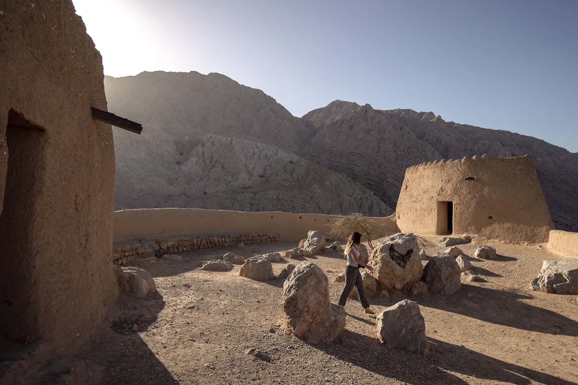 A woman walks within the twin turrets and wall fortifications of Dhayah Fort in the mountain backed desert landscape of Ras Al Khaimah.