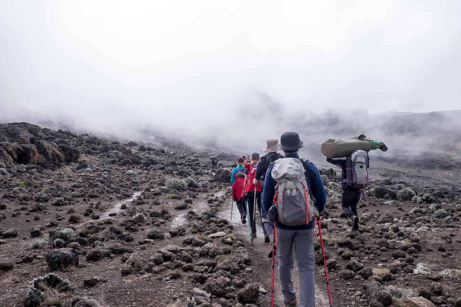 A porter on the right carrying a large, long bag, passes a group of trekkers to the right on a rocky, volcanic landscape of Mt. Kilimanjaro.