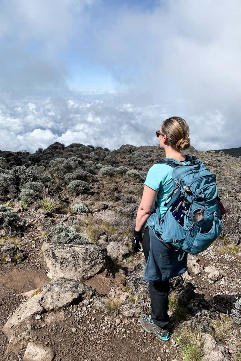 A woman in a blue t-shirt, with a blue daypack stands on a rocky path overlooking a Kilimanjaro valley covered in clouds.