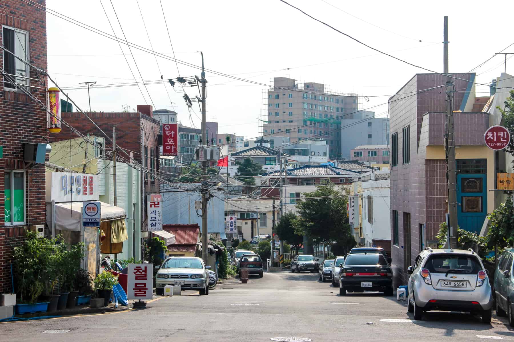 Low rise, pastel-coloured boxed buildings of a neighbourhood in Jeju Island's capital of Jeju City. Electrical wires can be seen hanging between the houses and a high rise complex can be seen in the background. 