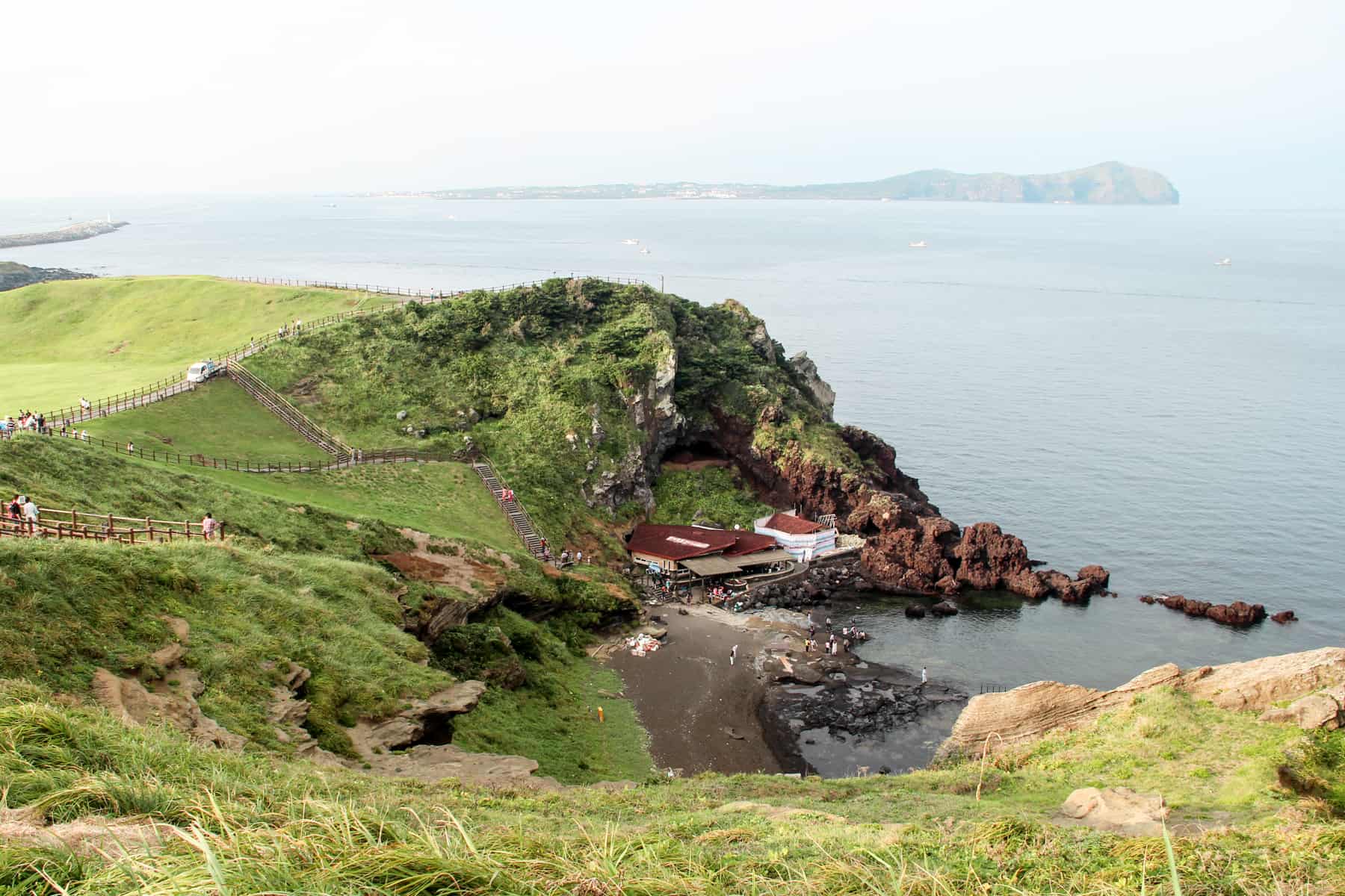People strolling up a series of narrow walking paths on a green mound of Jeju Island's Sunrise Peak, looking down onto red-roofed houses, coastal rocks and the vast ocean.