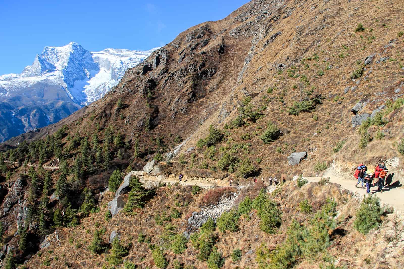 Four people trek up a long, narrow, sandy coloured mountain ridge pathway on the Everest Base Camp Trek. IN the background is the snow capped Himalaya peaks