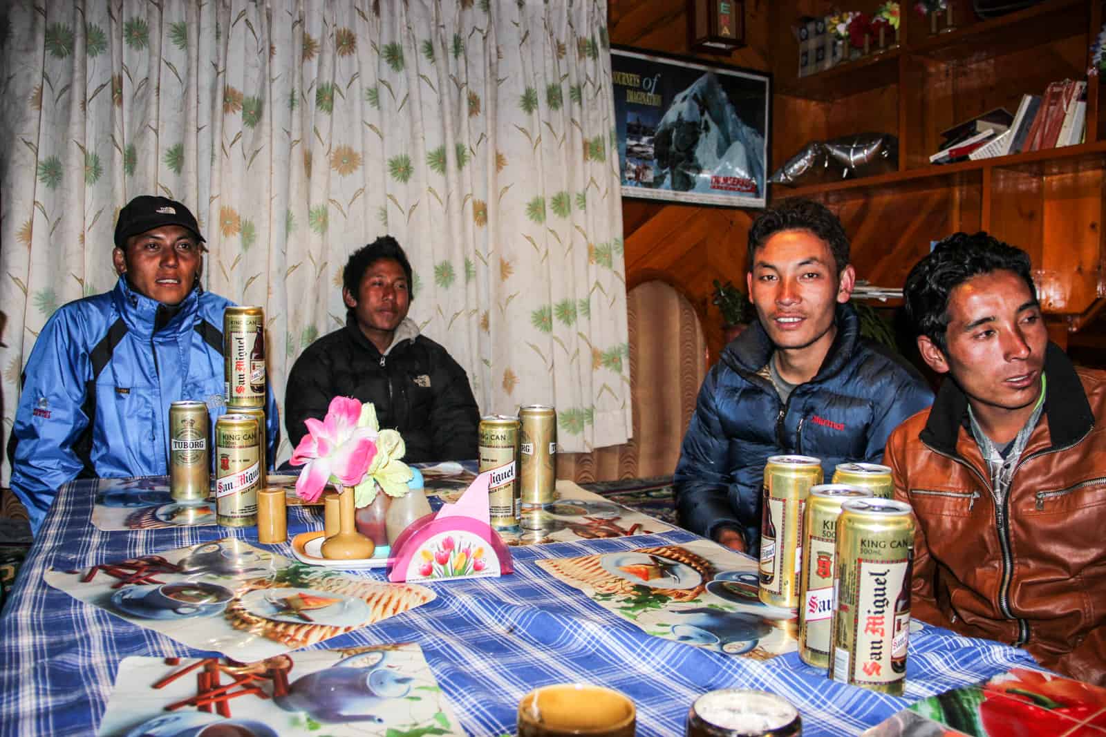 Four sherpas on the Everest Base Camp trek enjoy post-trek beers on a blue and white checkered table in a wooden mountain hut