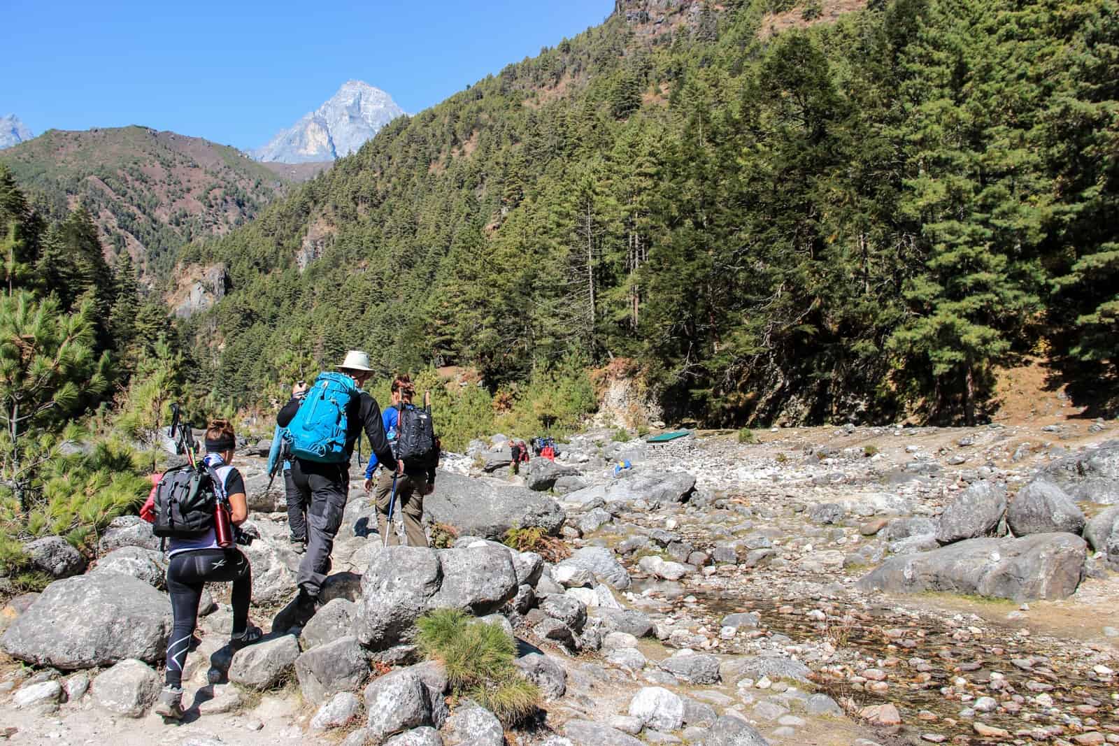 Three hikers cfoss a rocky scree backed by dense green forests and mountainous hills on the Everest Base Camp Trek in Nepal